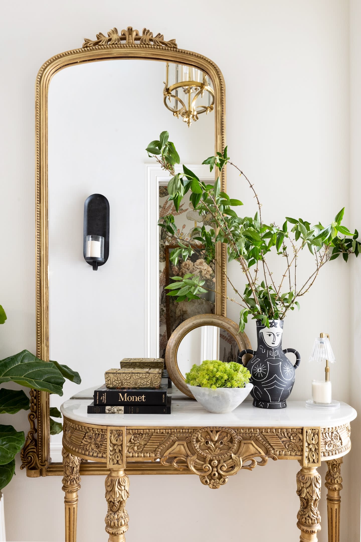 An ornate gold-framed mirror sits above an intricately carved table with a white top. On the table are books, a decorative vase, and plants. The setting feels elegant and classic.