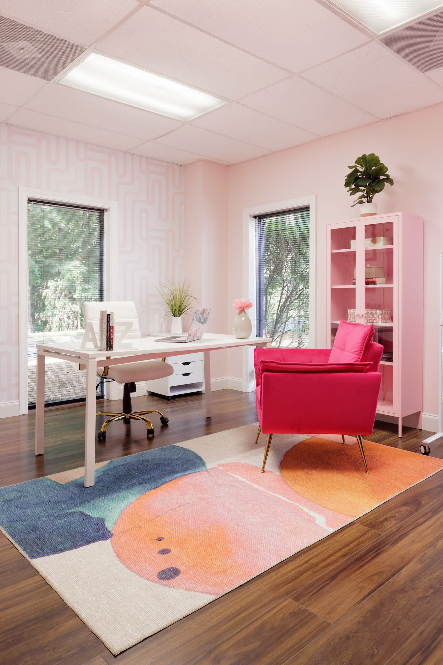 Bright office with pink walls, featuring a white desk, modern white chair, hot pink armchair, colorful abstract rug, and a pink cabinet, creating a lively and cheerful atmosphere.