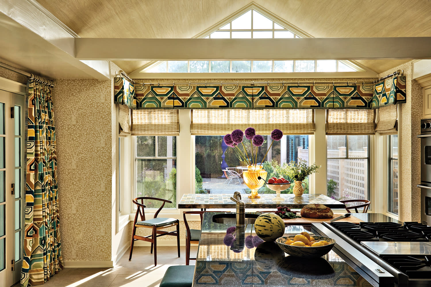 Bright kitchen with geometric-patterned curtains and Roman blinds. Sunlight filters through large windows, illuminating a table with flowers and fruit.