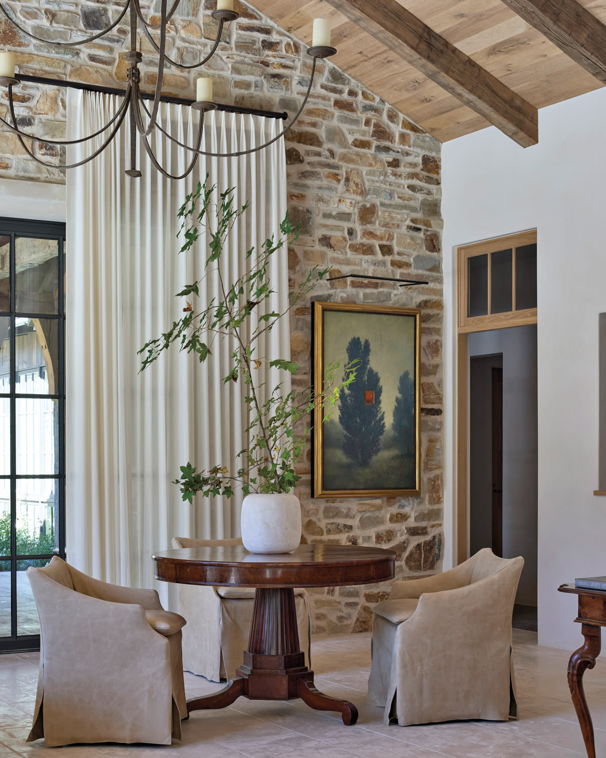 Elegant room with stone walls, wooden ceiling, and a chandelier. Features a round table, beige chairs, a tall plant, and framed artwork, conveying warmth.