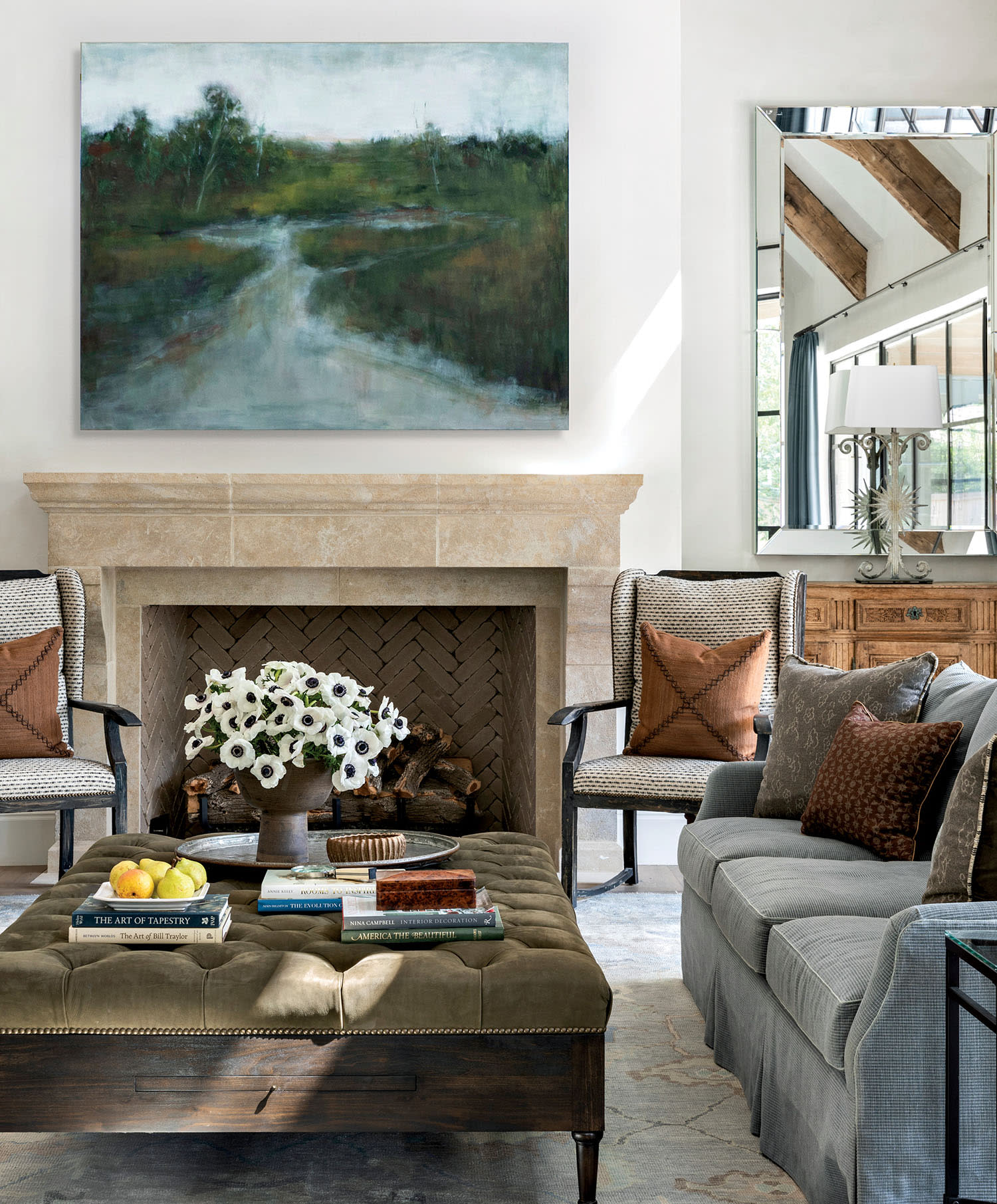 Elegant living room with a beige stone fireplace, nature painting above. Cozy armchairs with brown cushions, gray sofa, and tufted ottoman with books and flowers. Bright, calm ambiance.